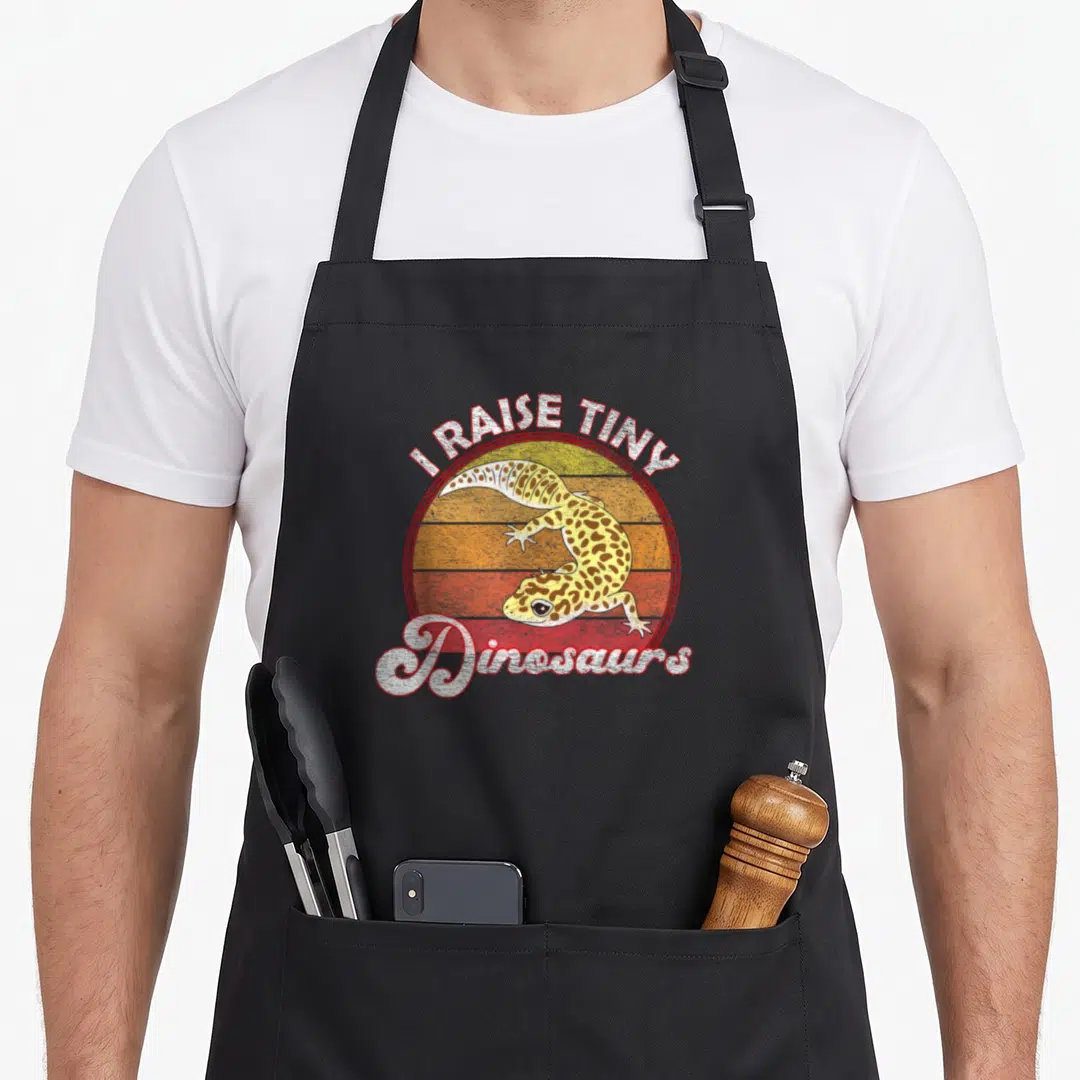 Dinosaur leopard apron - Retro sun graphic with leopard gecko and I RAISE TINY Dinosaurs text — shown on a black cotton canvas apron against a white background