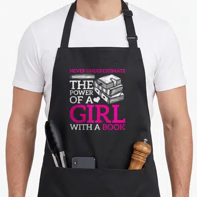 Underestimate reading book apron - Pink and white text reading Never Underestimate Power of Girl With Book over stacked — shown on a black cotton canvas apron against a white background