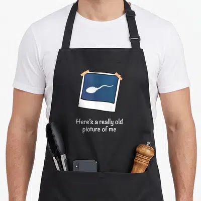 Funny sperm gag aprons - Cartoon polaroid of white sperm on blue background with orange tape and text below — shown on a black cotton canvas apron against a white background
