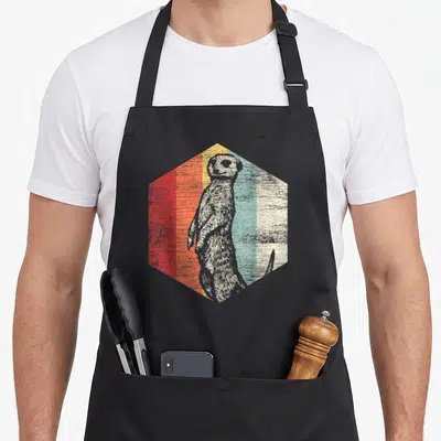 Retro meerkat apron - Grey meerkat inside hexagon with red orange yellow blue cream vertical stripes distressed — shown on a black cotton canvas apron against a white background