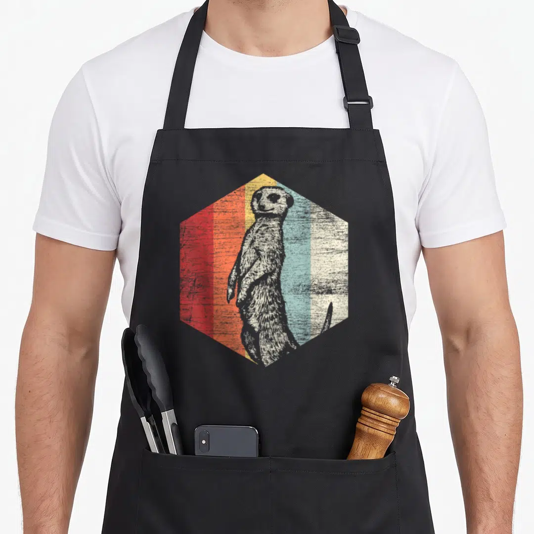 Retro meerkat apron - Grey meerkat inside hexagon with red orange yellow blue cream vertical stripes distressed — shown on a black cotton canvas apron against a white background