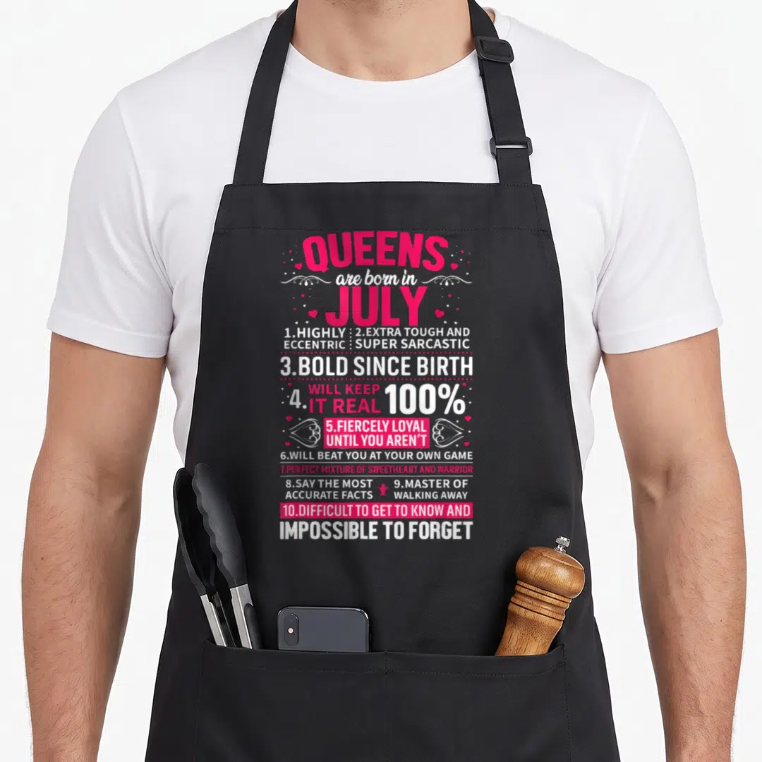 July queen apron - Queens are born in July typography with ten personality trait lines and small hearts — shown on a black cotton canvas apron against a white background