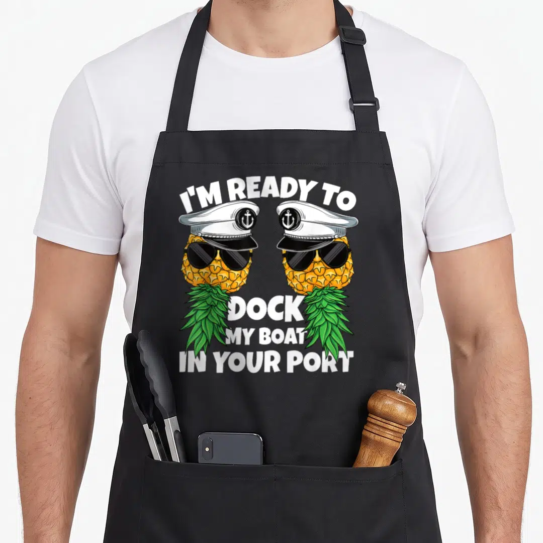 Ready dock apron - Two cartoon pineapples in captain hats and sunglasses with arched nautical text — shown on a black cotton canvas apron against a white background