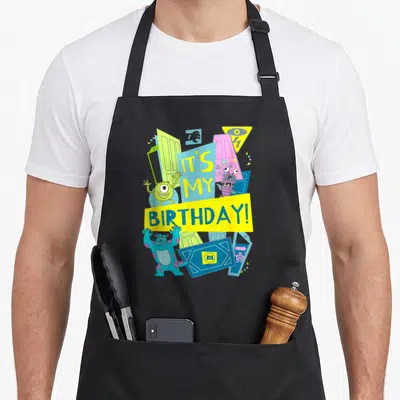 Cool birthday apron for baking - Cartoon monsters and tilted doors surround IT'S MY BIRTHDAY! text on yellow and blue — shown on a black cotton canvas apron against a white background