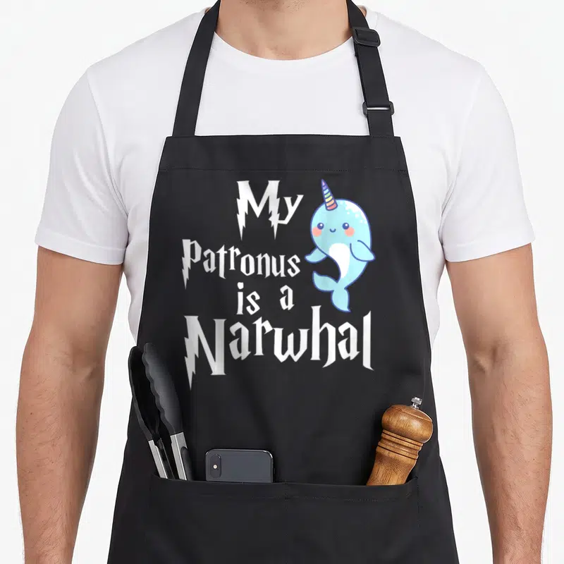 Patronus narwhal apron - Stacked white text My Patronus is a Narwhal beside light blue cartoon narwhal with rainbow horn — shown on a black cotton canvas apron against a white background