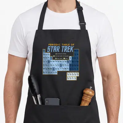Map table apron - Star Trek periodic table grid with yellow blue grey white cells listing captains crew villains — shown on a black cotton canvas apron against a white background