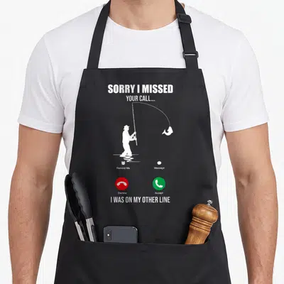 Sorry missed fishing aprons - Fisherman silhouette with phone interface icons and text SORRY I MISSED YOUR CALL — shown on a black cotton canvas apron against a white background