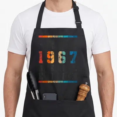 Limited special aprons - Retro 1967 text print with red orange and blue blocks on grey background — shown on a black cotton canvas apron against a white background