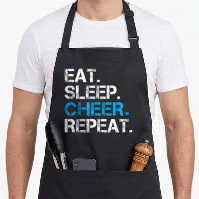 Eat rally camping apron - Stacked EAT SLEEP CHEER REPEAT text in distressed white and blue with a small heart next — shown on a black cotton canvas apron against a white background