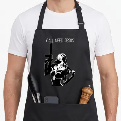 Nun machine apron - White Y'all Need Jesus text above black and white line art of a masked nun holding a rifle — shown on a black cotton canvas apron against a white background
