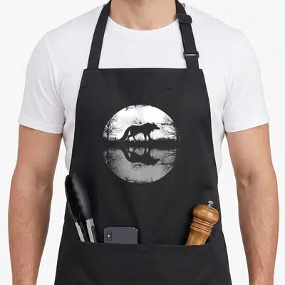 Moon wolf apron - Grey wolf silhouette walking near trees inside a white moon circle with water reflection below — shown on a black cotton canvas apron against a white background