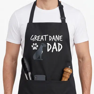 Dane puppy dog apron - Grey cartoon Great Dane sitting between white paw print and text GREAT DANE DAD — shown on a black cotton canvas apron against a white background