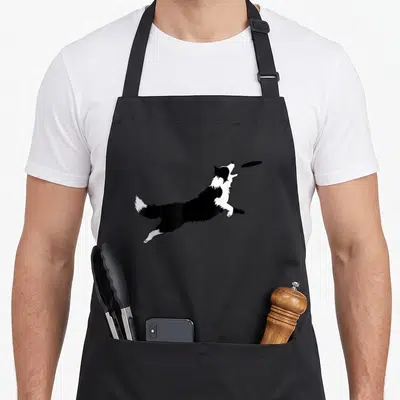 Fetch border collie apron - Black and white dog leaping to catch a flying disc — shown on a black cotton canvas apron against a white background