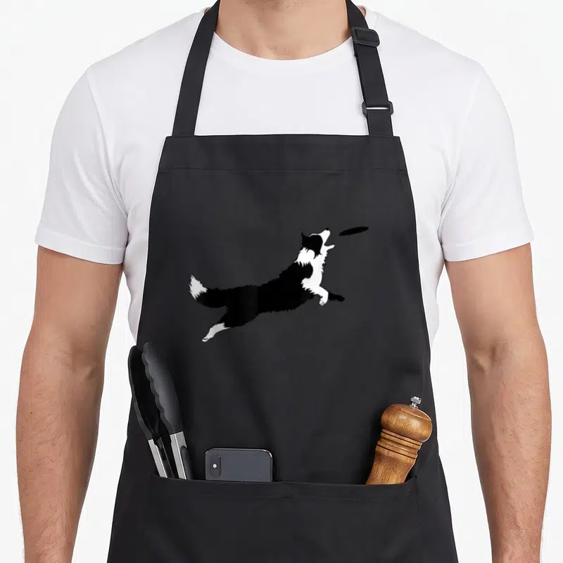Fetch border collie apron - Black and white dog leaping to catch a flying disc — shown on a black cotton canvas apron against a white background