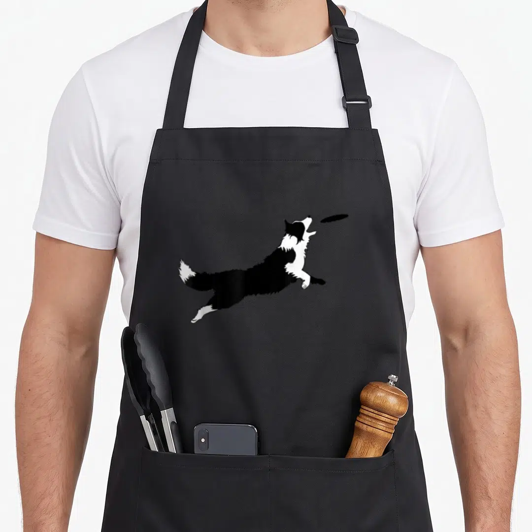 Fetch border collie apron - Black and white dog leaping to catch a flying disc — shown on a black cotton canvas apron against a white background