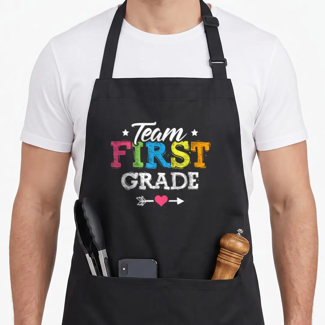 Grade student teacher apron - Stacked Team First Grade text in rainbow colors with arrow and pink heart below — shown on a black cotton canvas apron against a white background