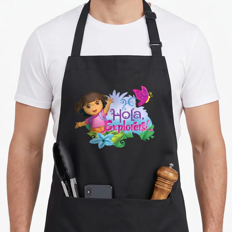 Dora spring apron - Dora waving next to Hola Explorers text with butterfly and blue flower — shown on a black cotton canvas apron against a white background