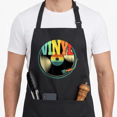 Record collector vinyl apron - Rainbow VINYL text over a black record with a tonearm in retro colors — shown on a black cotton canvas apron against a white background
