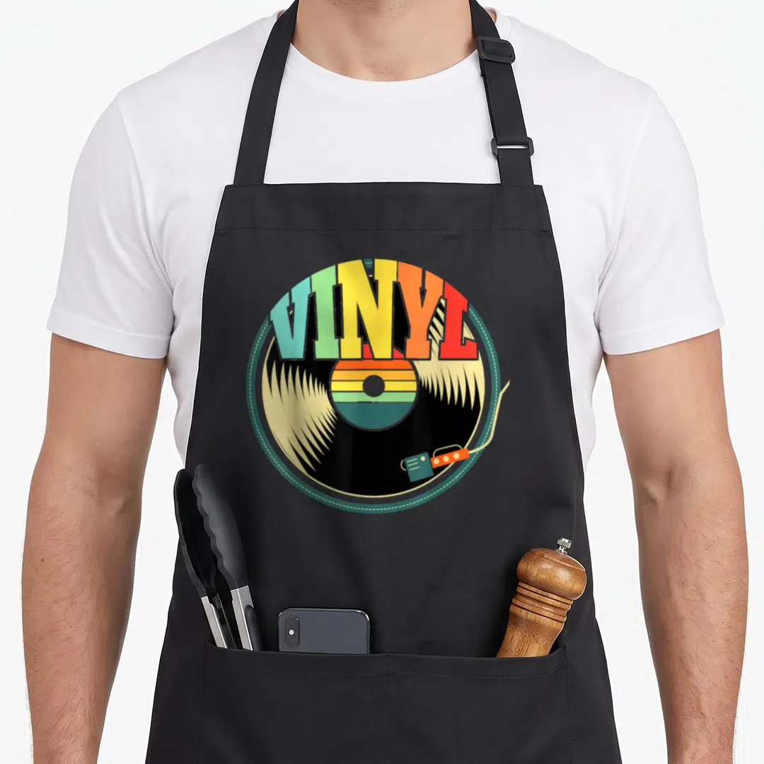 Record collector vinyl apron - Rainbow VINYL text over a black record with a tonearm in retro colors — shown on a black cotton canvas apron against a white background