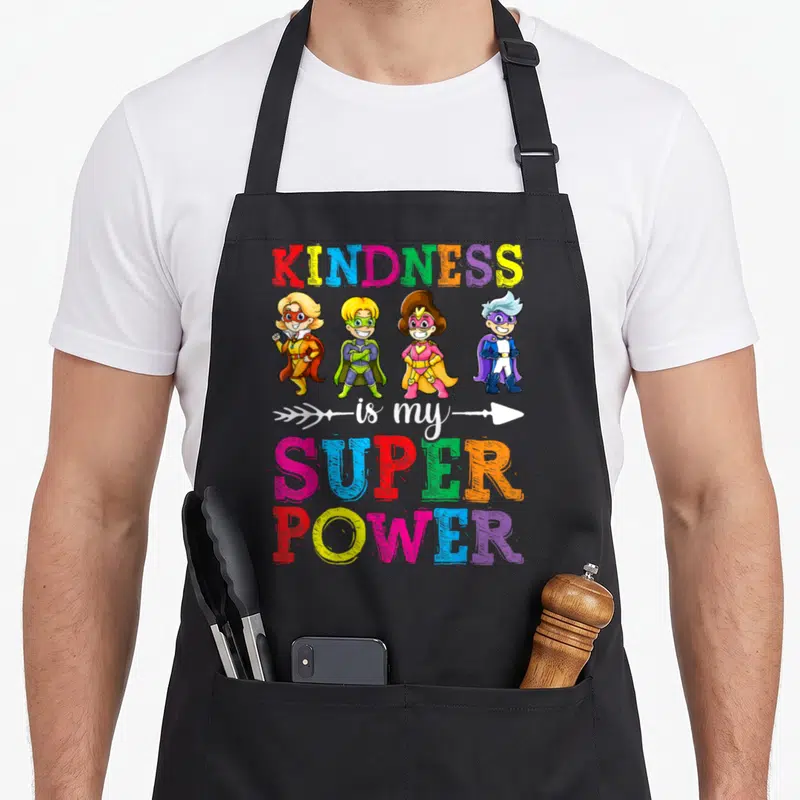 Superpower child kindness apron - Four cartoon superheroes below rainbow text reading KINDNESS is my SUPER POWER — shown on a black cotton canvas apron against a white background