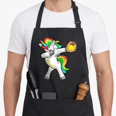 Softball rainbow apron - Cartoon white unicorn dabbing in brown glove holding yellow softball — shown on a black cotton canvas apron against a white background