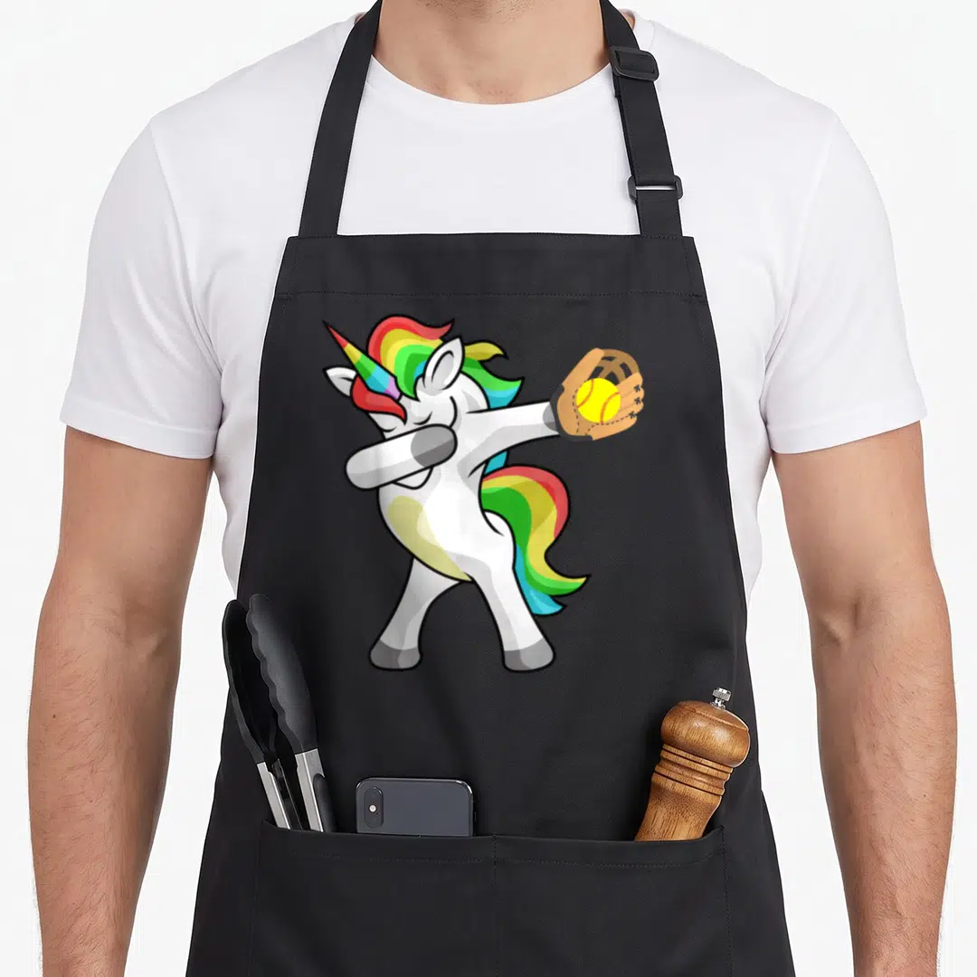 Softball rainbow apron - Cartoon white unicorn dabbing in brown glove holding yellow softball — shown on a black cotton canvas apron against a white background