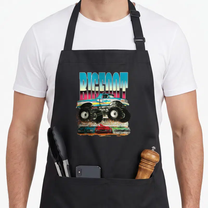 Wildfoot car apron - Retro monster truck crushing three cars under BIGFOOT text in blue pink red green — shown on a black cotton canvas apron against a white background