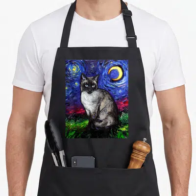 Starry cute cat aprons - Siamese cat sitting on green grass under a swirling blue night sky with a yellow moon — shown on a black cotton canvas apron against a white background
