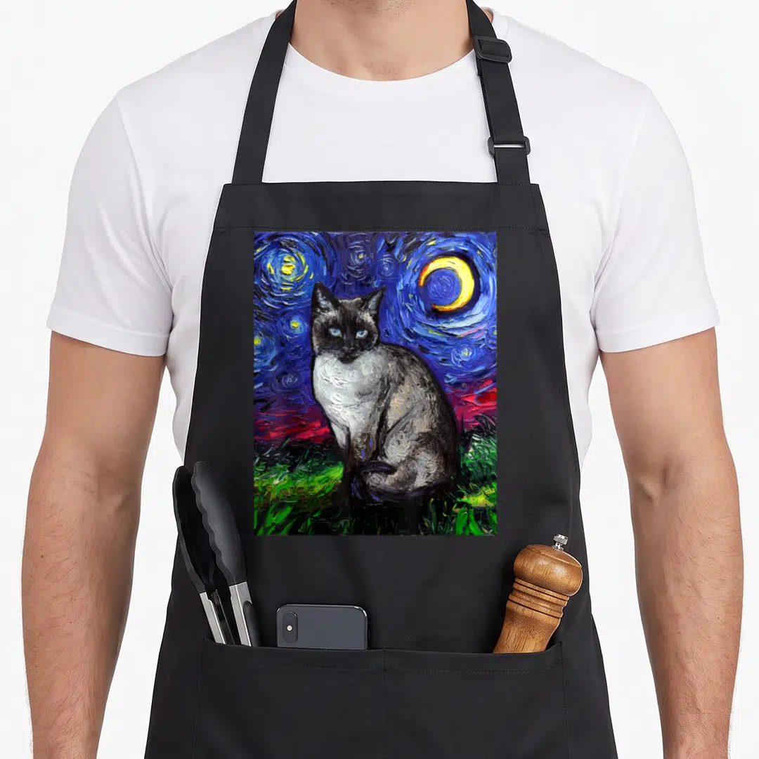 Starry cute cat aprons - Siamese cat sitting on green grass under a swirling blue night sky with a yellow moon — shown on a black cotton canvas apron against a white background