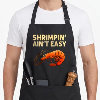 Easy fishing aprons - Beige text Shrimpin Ain Easy above orange cartoon shrimp — shown on a black cotton canvas apron against a white background