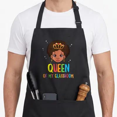 Funny queen classroom aprons - Cartoon girl with brown skin and gold crown above multi-colored QUEEN OF MY CLASSROOM — shown on a black cotton canvas apron against a white background