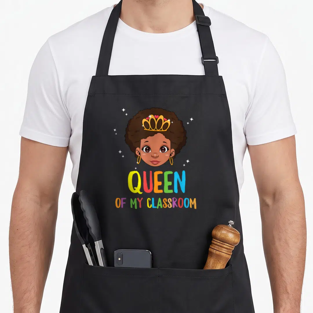 Funny queen classroom aprons - Cartoon girl with brown skin and gold crown above multi-colored QUEEN OF MY CLASSROOM — shown on a black cotton canvas apron against a white background