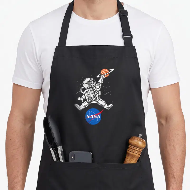Basketball nasa apron - Line art astronaut holding orange ringed planet above blue red NASA logo — shown on a black cotton canvas apron against a white background