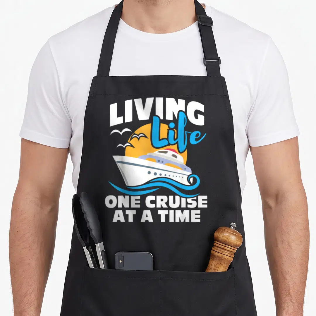 Living cruiser apron - White cruise ship on blue waves under yellow sun with LIVING Life ONE CRUISE AT A TIME text — shown on a black cotton canvas apron against a white background