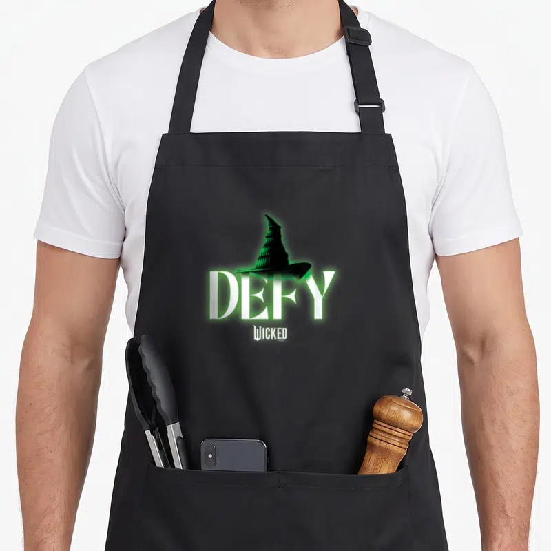 Wicked tie apron - Green and black witch hat above white DEFY text and small Wicked logo — shown on a black cotton canvas apron against a white background
