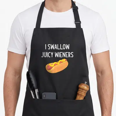 Swallow joke apron - I Swallow Juicy Wieners text above cartoon hot dog with mustard — shown on a black cotton canvas apron against a white background