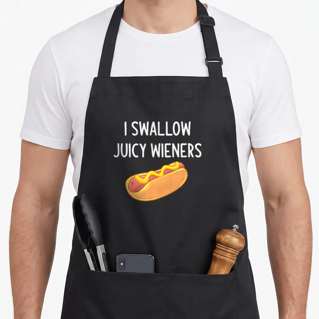 Swallow joke apron - I Swallow Juicy Wieners text above cartoon hot dog with mustard — shown on a black cotton canvas apron against a white background