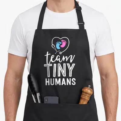Neonatal tiny chef apron - Stethoscope heart with blue and pink footprints above stacked white text reading team TINY — shown on a black cotton canvas apron against a white background