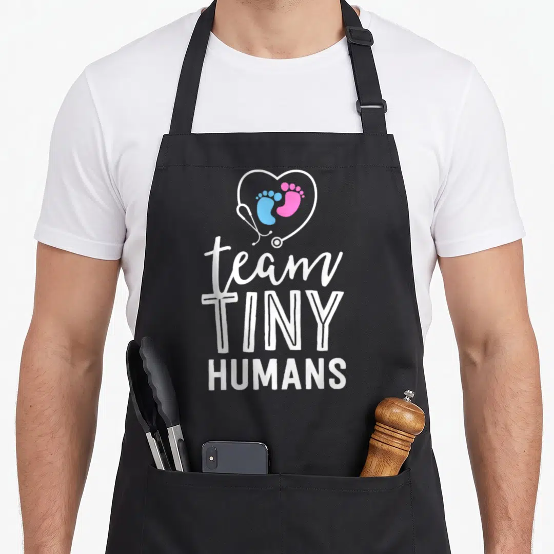 Neonatal tiny chef apron - Stethoscope heart with blue and pink footprints above stacked white text reading team TINY — shown on a black cotton canvas apron against a white background