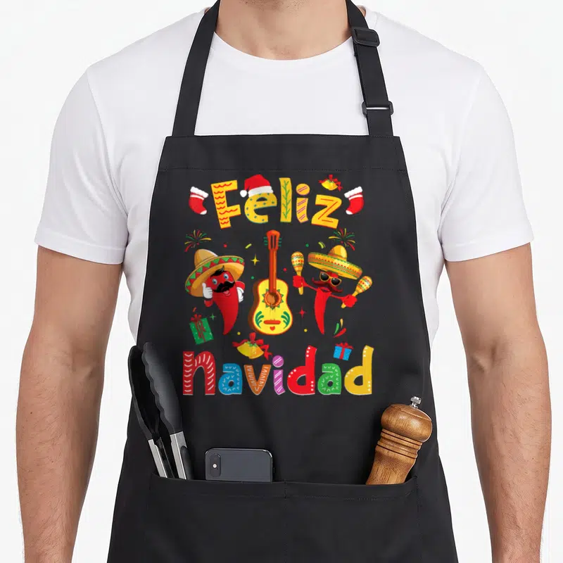 Navidad xmas apron - Two chili peppers in sombreros flank a guitar under Feliz navidad text in red yellow green blue — shown on a black cotton canvas apron against a white background