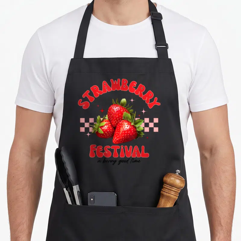 Festival strawberry apron - Three red strawberries over pink checkerboard with STRAWBERRY FESTIVAL text and a berry — shown on a black cotton canvas apron against a white background