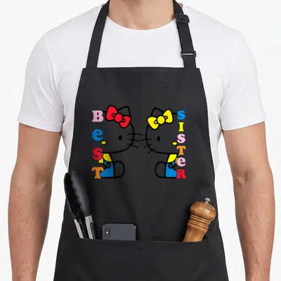 Sister hello apron - Two Hello Kitty figures face each other with vertical BEST SISTER text in pink, blue, red, yellow — shown on a black cotton canvas apron against a white background