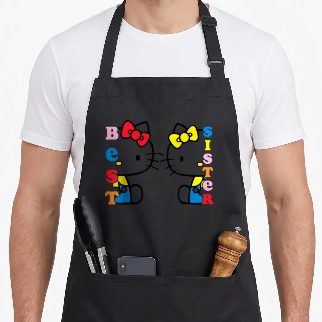 Sister hello apron - Two Hello Kitty figures face each other with vertical BEST SISTER text in pink, blue, red, yellow — shown on a black cotton canvas apron against a white background