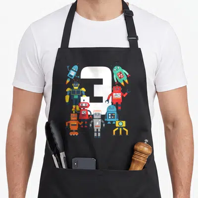 Robotic science apron - White number 3 surrounded by eight colorful cartoon robots in red blue green yellow grey — shown on a black cotton canvas apron against a white background