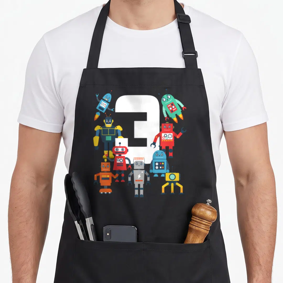 Robotic science apron - White number 3 surrounded by eight colorful cartoon robots in red blue green yellow grey — shown on a black cotton canvas apron against a white background