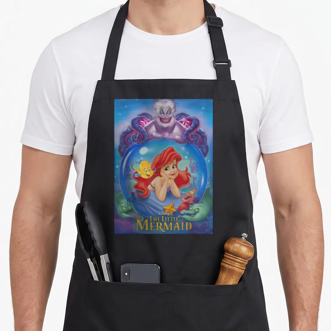 Ariel ursula mermaid apron - Ursula looming over bubble with Ariel and Flounder, Sebastian and star below, Disney — shown on a black cotton canvas apron against a white background
