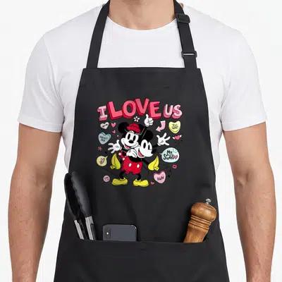 Valentine's day apron gift - Mickey and Minnie hugging under I LOVE US text with hearts and sweet phrases — shown on a black cotton canvas apron against a white background