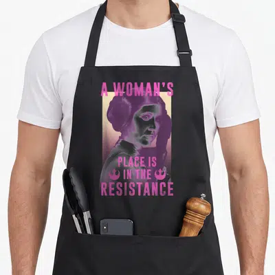 Resistance princess apron - Retro portrait of a woman holding a blaster with pink text reading A Woman's Place Is — shown on a black cotton canvas apron against a white background