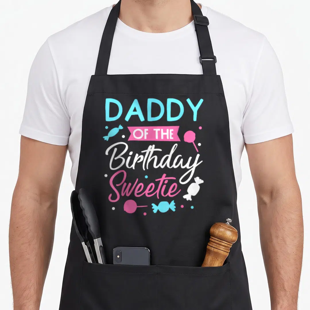 Sweetie daddy apron - DADDY OF THE Birthday Sweetie text with cyan and pink candies and lollipops — shown on a black cotton canvas apron against a white background