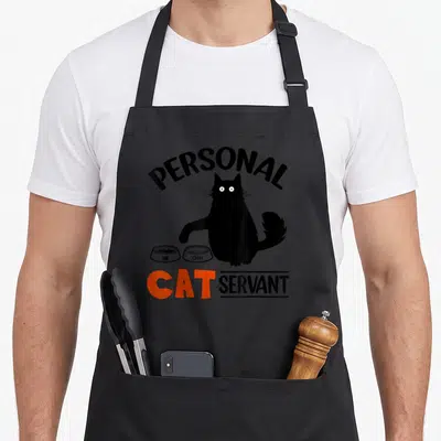 Cat personal apron - Black cat sitting between two bowls with arched and stacked text in black and orange — shown on a black cotton canvas apron against a white background